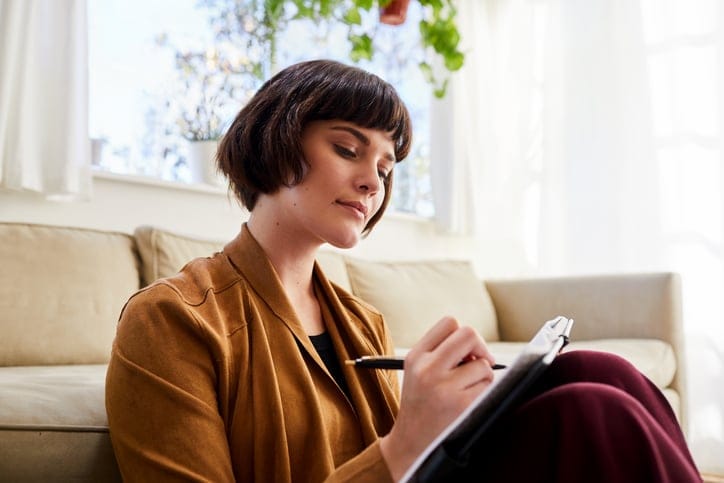 A young woman sitting near the couch writing notes or journaling.