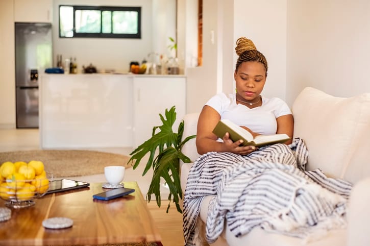 Young woman reading book under blanket on couch.
