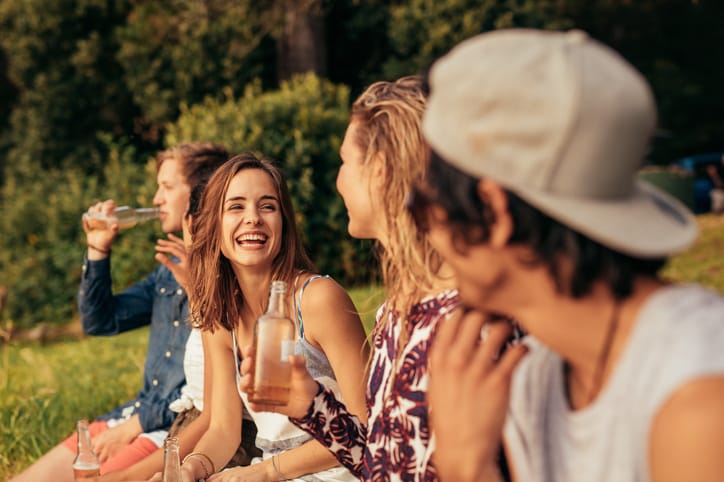 Portrait of happy young people sitting by a lake with beers. Group of friends hanging out at the lake.