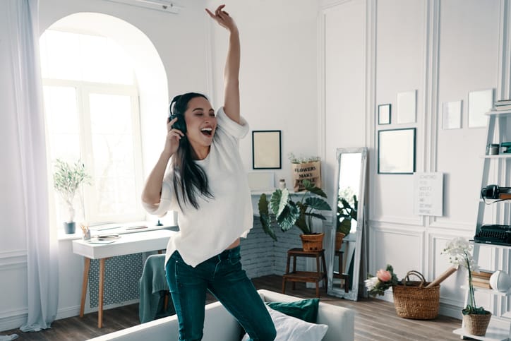Full length of attractive young woman wearing headphones and smiling while dancing on sofa at home