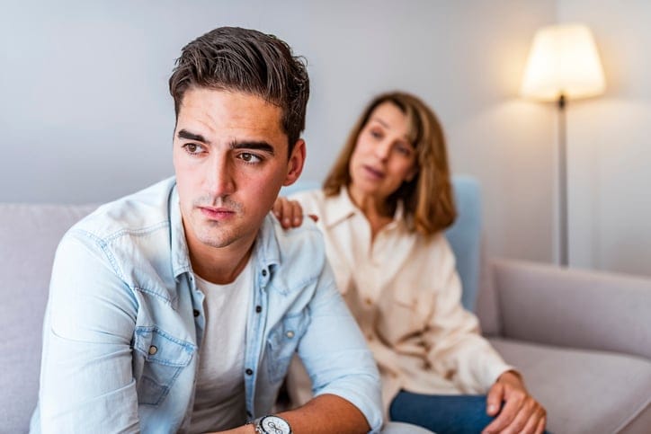 Mature Mother and problem child. Frustrated mother talking to with her upset son sitting on the couch. Senior mother talking with her unhappy son at home during the day. Mother comforting her black hair sad son.