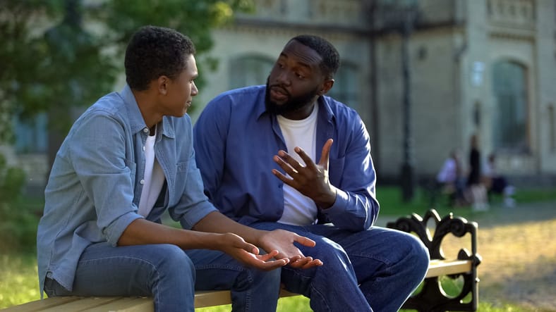 Father arguing teenage son sitting on campus bench, puberty age difficulties