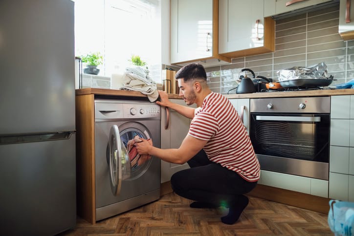 Man crouching down to put washing into his washing machine in his kitchen. He is in the Northeast of England.