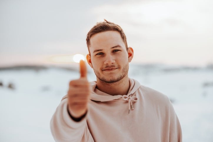 Smiling man standing in snow covered winter landscape showing a thumbs up.