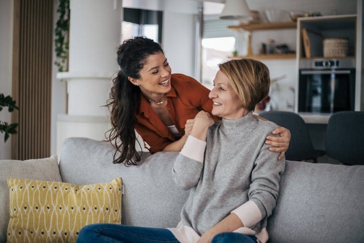 Smiling young woman hugging her mother who is sitting on sofa from behind.