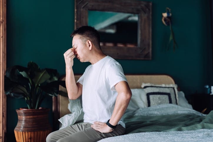 Senior Asian man with eyes closed feeling sick and discomfort, suffering from headache while sitting on bed at home. Elderly and health issues concept