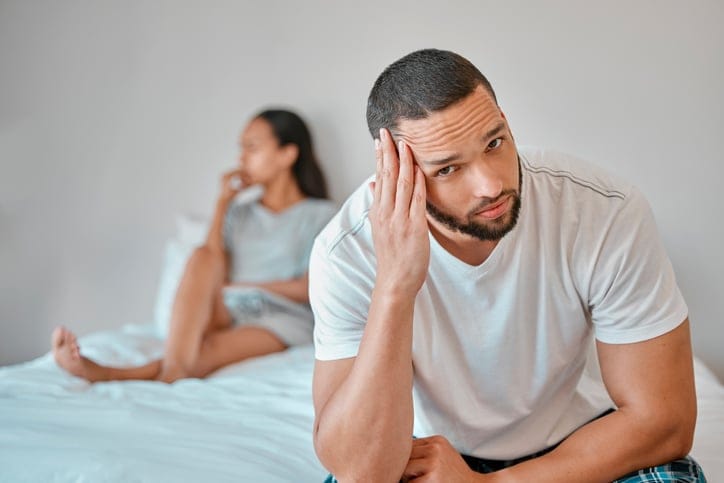 man upset on end of bed, girlfriend behind