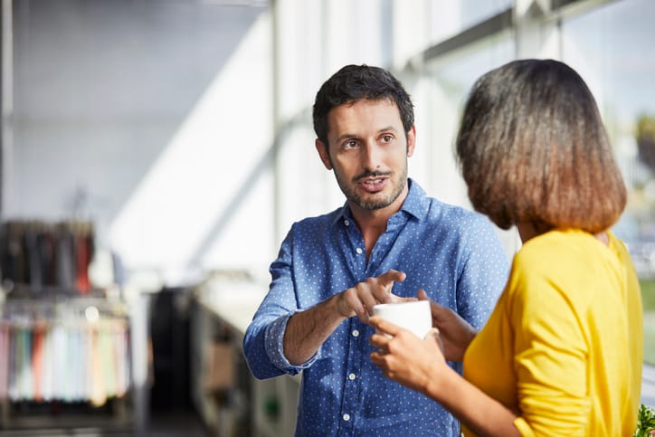 Confident businessman communicating with colleague having coffee. Male and female professionals are standing in office. They are discussing strategies at textile industry.