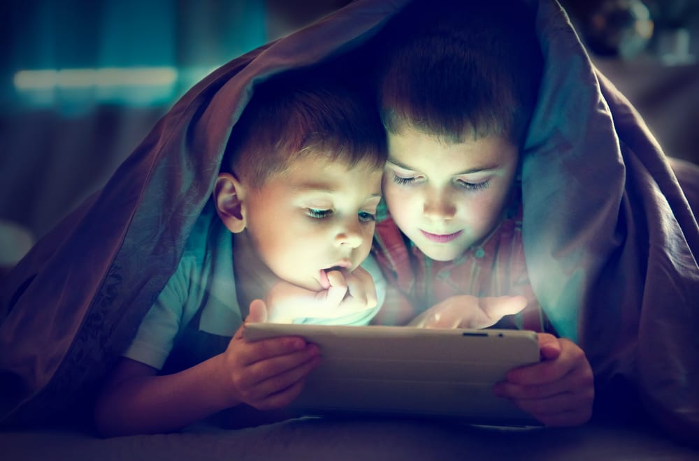 Two boys under a blanket at night reading on a tablet.