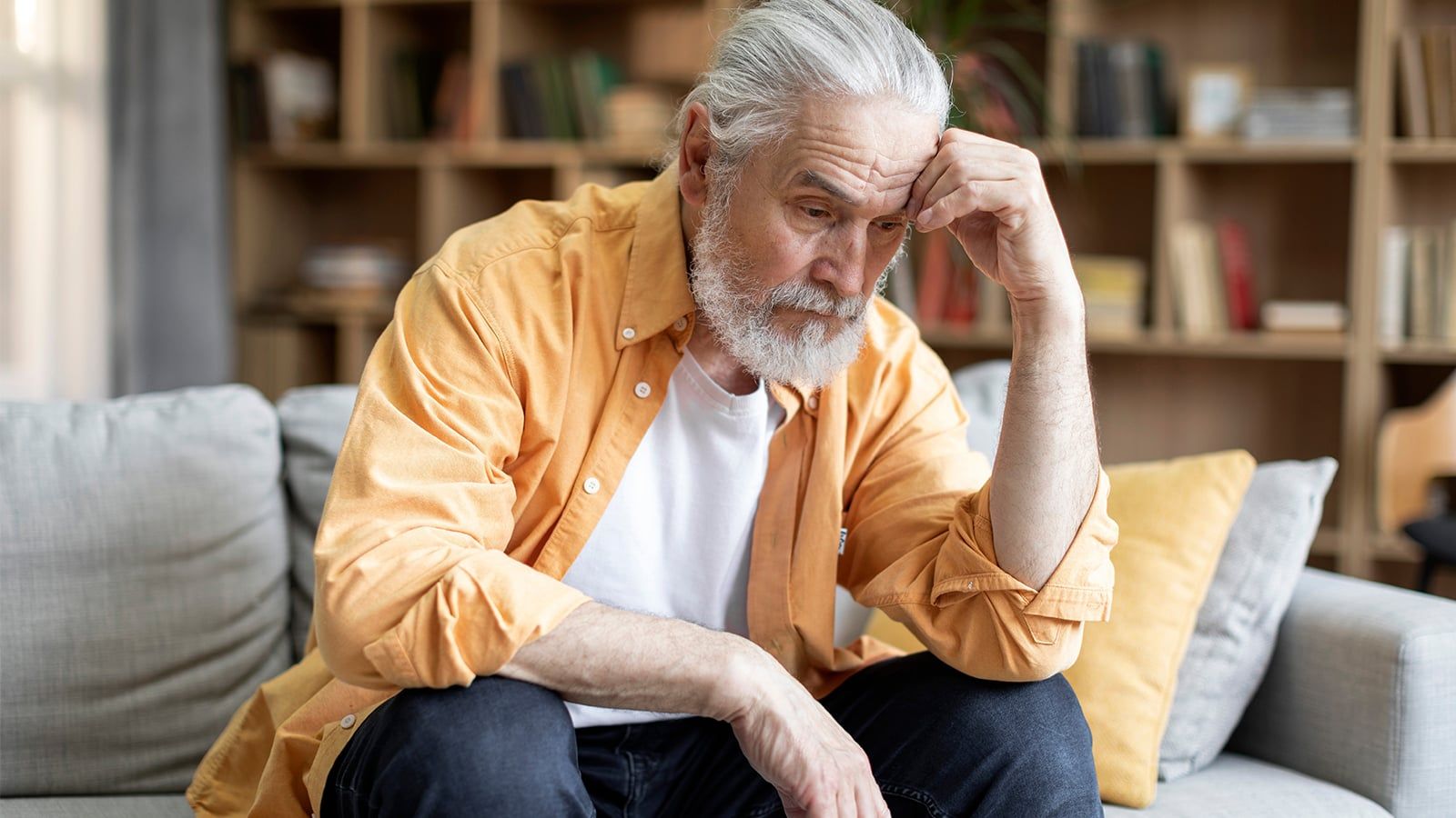 Upset bearded grey-haired old man in casual outfit sitting on couch alone, touching his head, looking down, feeling lonely, having heavy thoughts, home interior, copy space