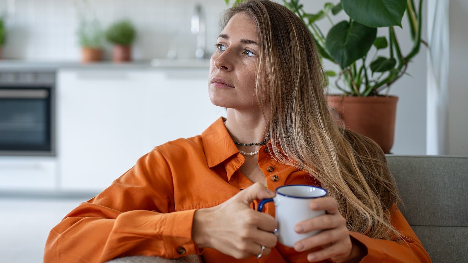 Thoughtful middle aged woman sits on sofa at home holding cup in hand looks towards window daydreaming. Peaceful pensive female thinking about future in cozy apartment in moment of quiet introspection