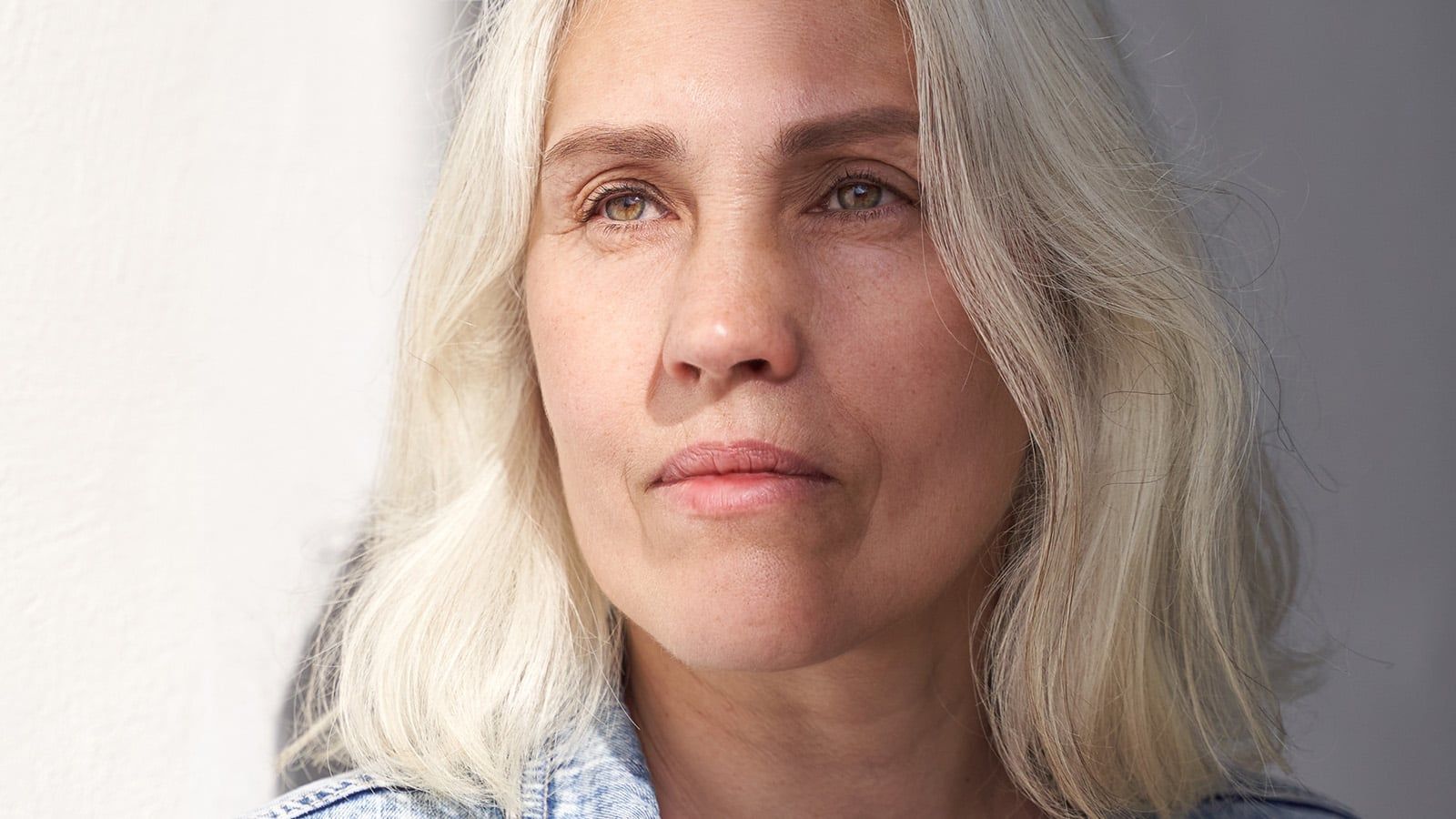 Mature woman with gray hair in relaxed denim outfit reflecting in natural light against minimalist background.