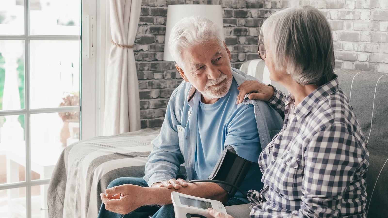 Senior woman using medical device to measure blood pressure of her husband suffering of hypertension - elderly couple sitting at home on sofa takes care of health