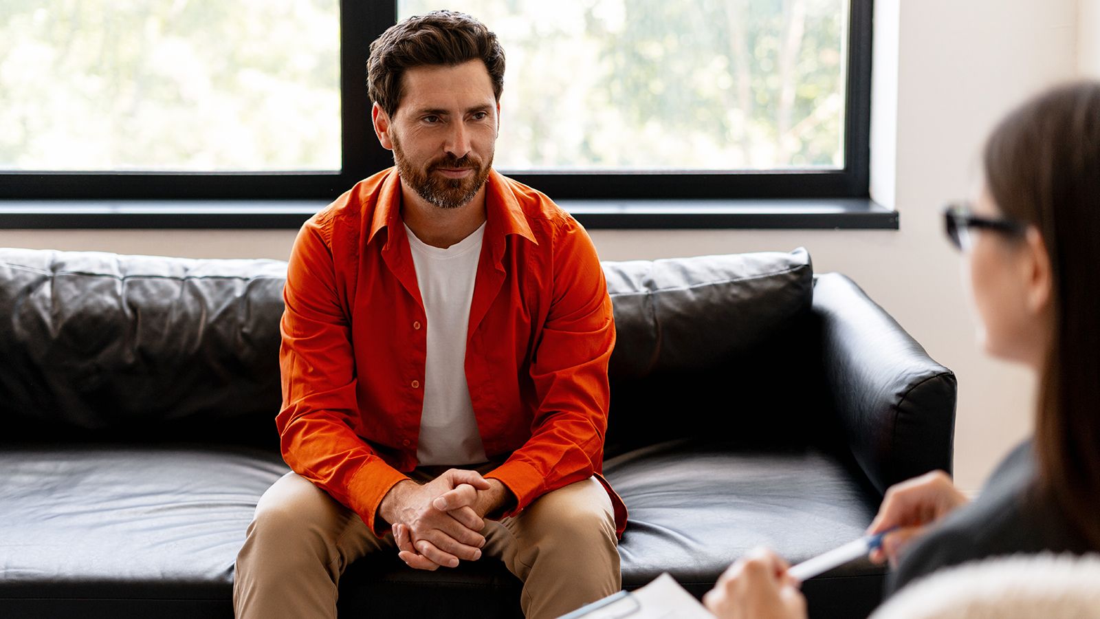 Bearded, middle aged man talking to psychologist during therapy session in modern office. Handsome male patient wearing casual red shirt.