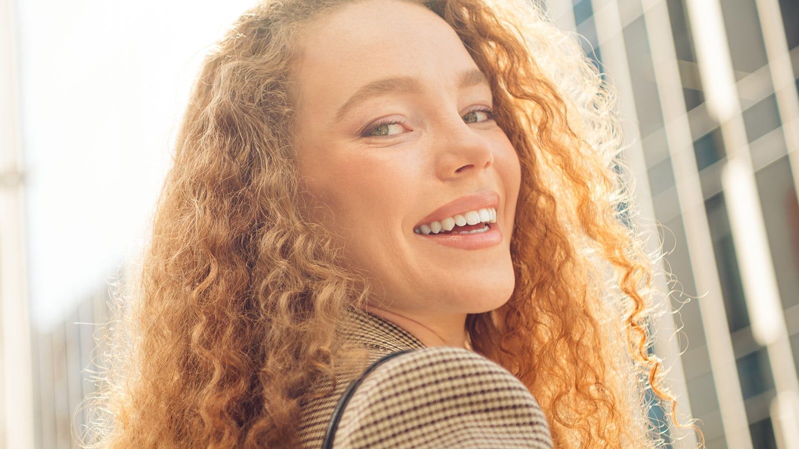Young professional woman with red curly hair smiling outdoors in a modern cityscape, exuding confidence and happiness