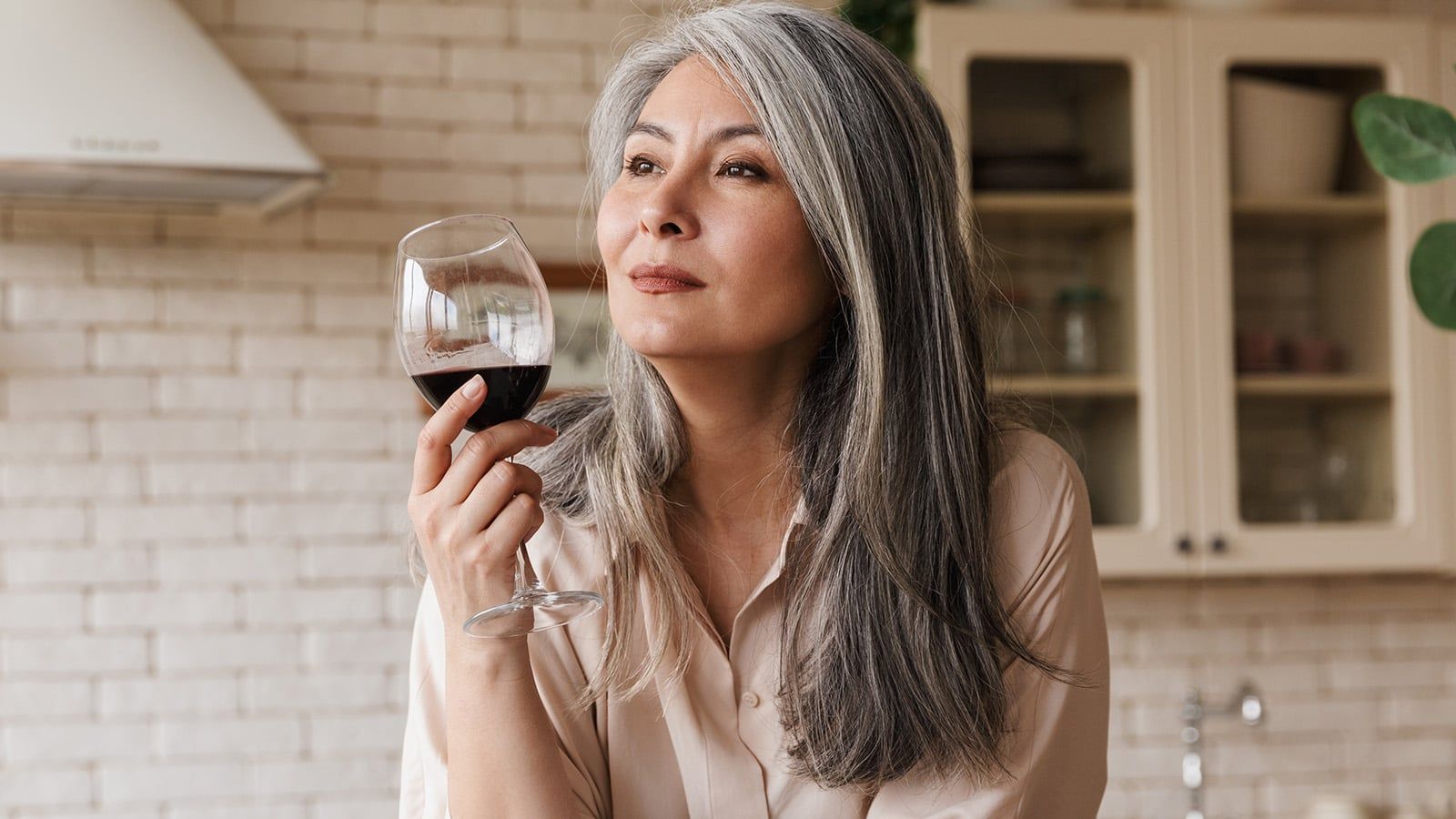 Picture of mature pretty woman sitting at the kitchen indoors at home drinking wine