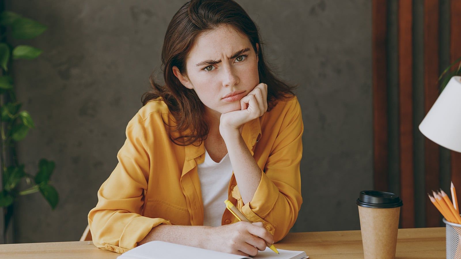 Young minded pensive thoughtful puzzled sad employee business woman 20s she wearing casual yellow shirt writing in notebook look camera sitting work at wooden office desk