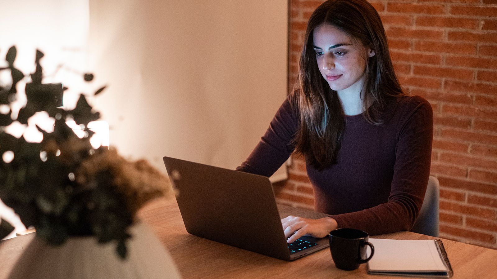 Concentrated young woman working late in home office, using laptop, copy space. Casual girl in the light of the screen on the background of the illuminated room. Technology and work concept