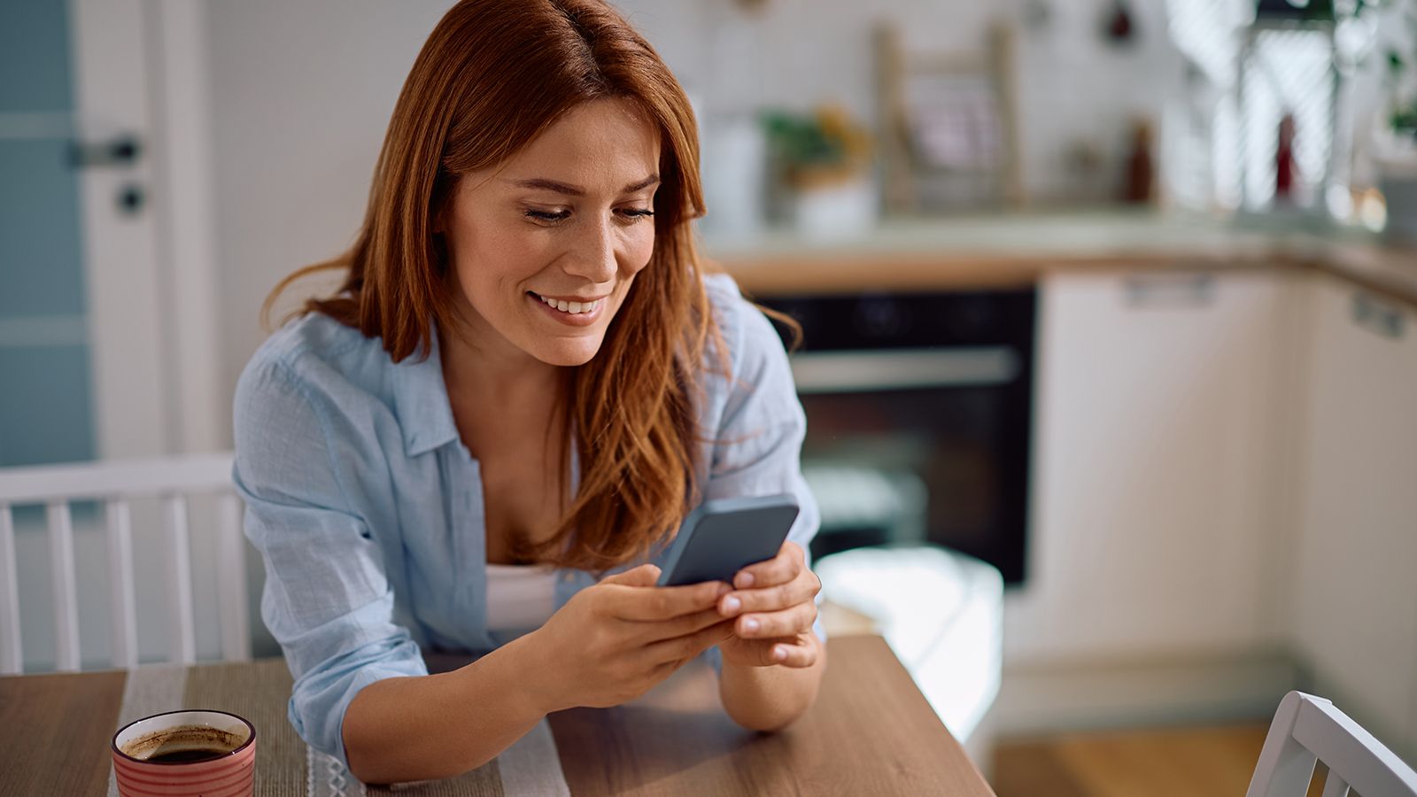 Happy woman texting on cell phone while drinking coffee at home.