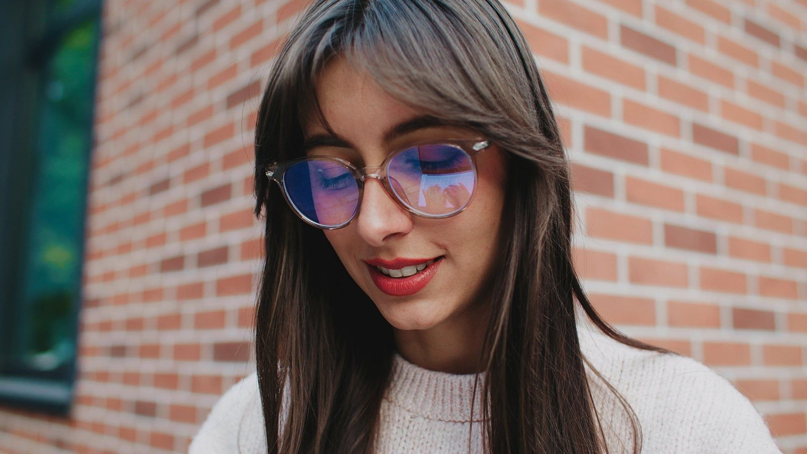 Close facial view of Caucasian girl with round glasses for better vision. Female looking at screen. Studying on digital device in fresh air. Beautiful long brown hair. Smiling to herself.