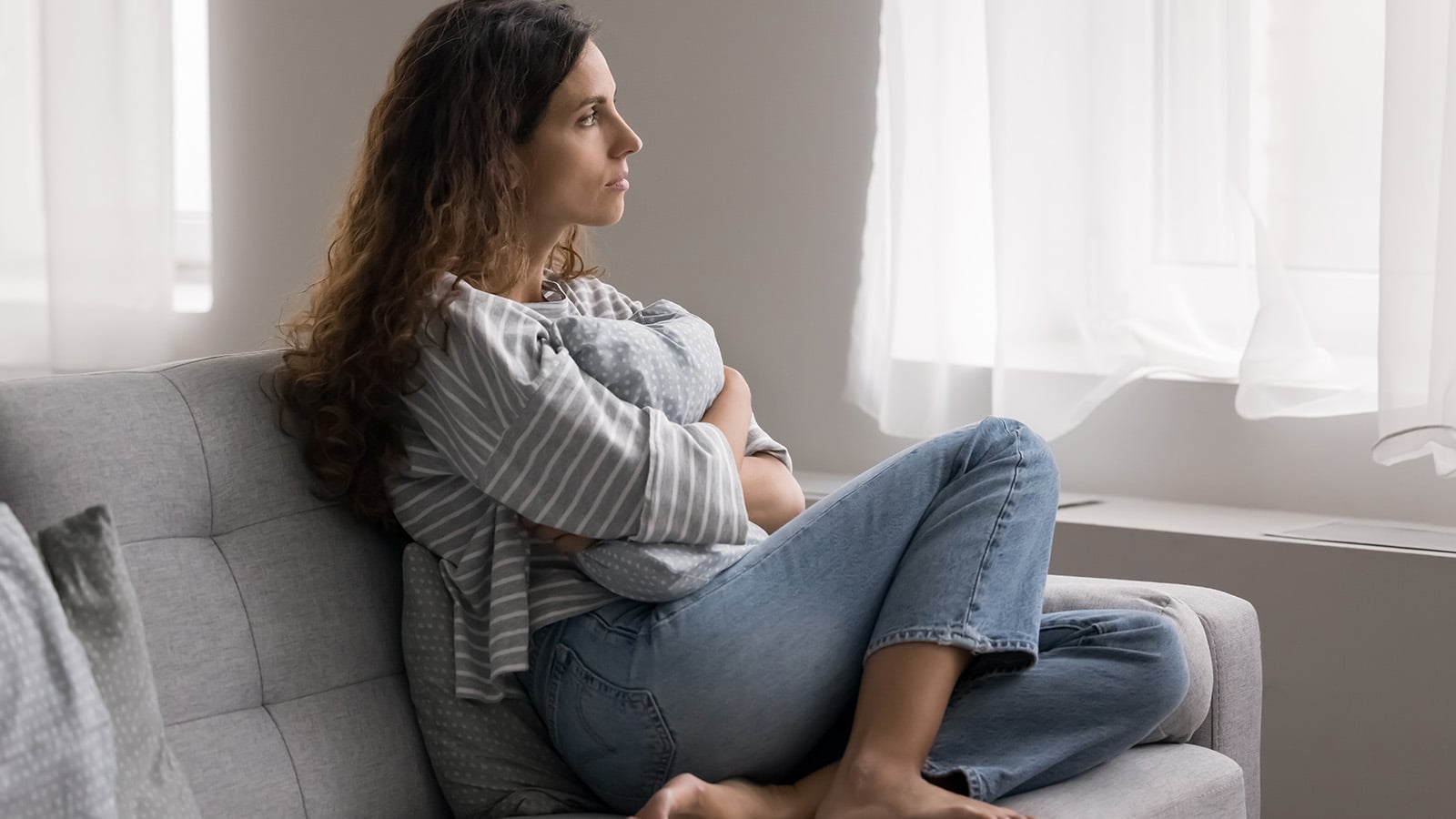 A woman sits on a couch, holding a baby wrapped in a blanket, and gazes thoughtfully out a bright window. She appears calm and pensive, dressed in a striped shirt and jeans in a softly lit room.