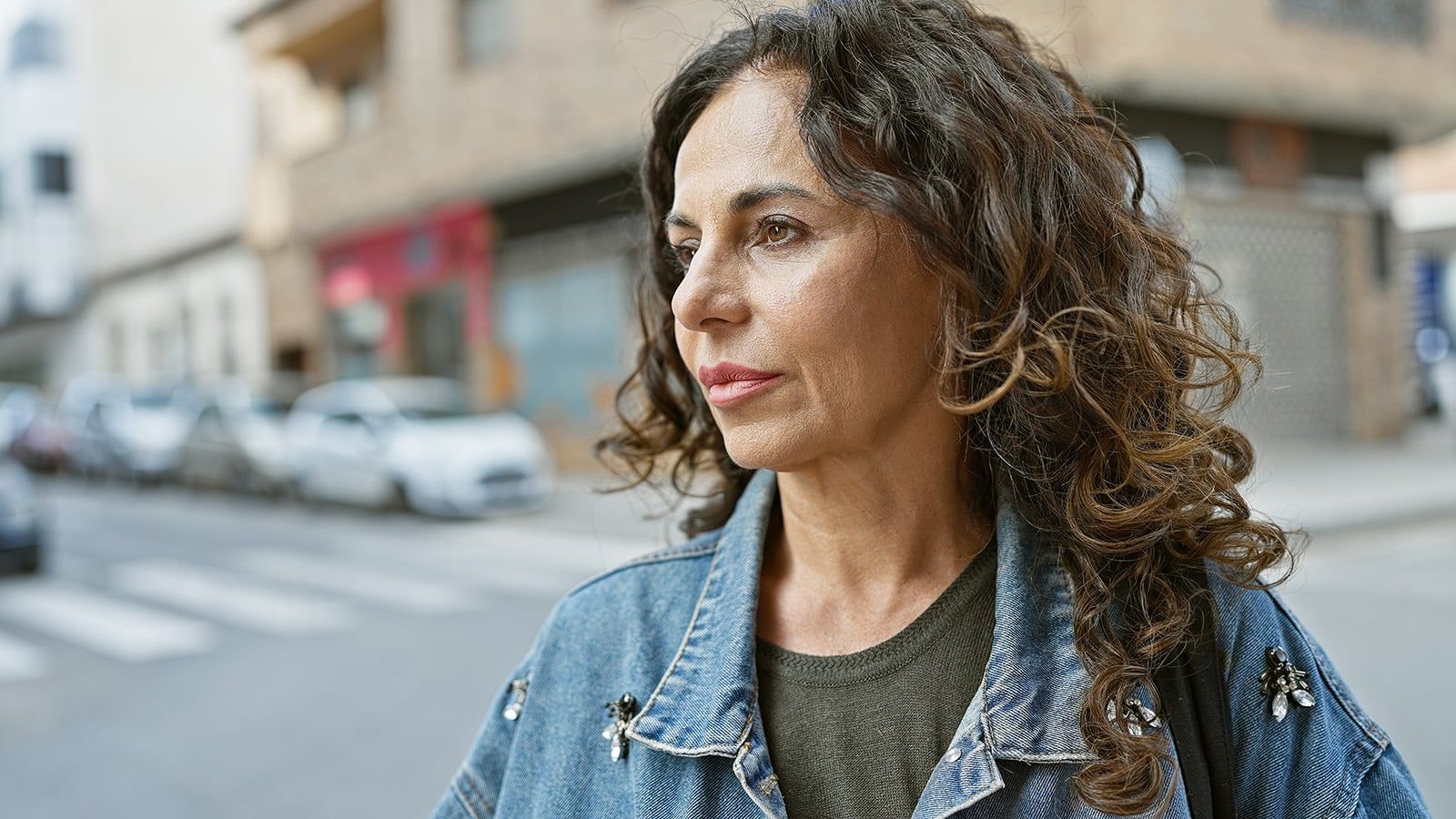 A woman with curly brown hair wearing a denim jacket stands outdoors on a city street, looking thoughtfully into the distance. Several parked cars and buildings are visible in the background.