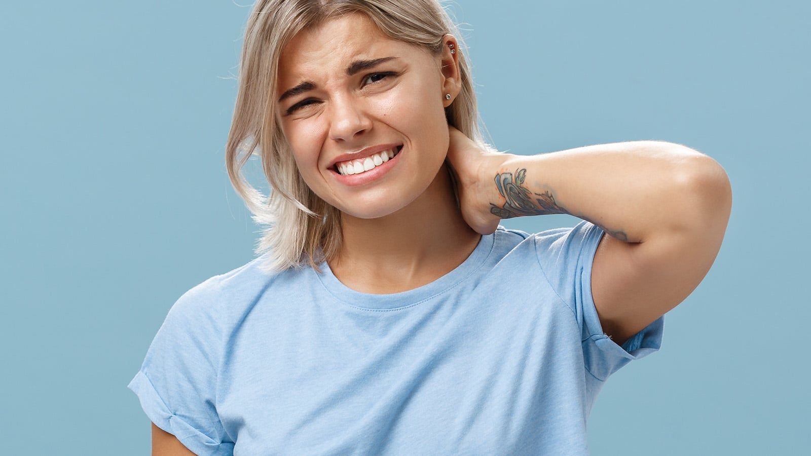 A young woman with short blonde hair wearing a light blue t-shirt looks uncomfortable and holds her neck in pain, grimacing against a plain blue background.