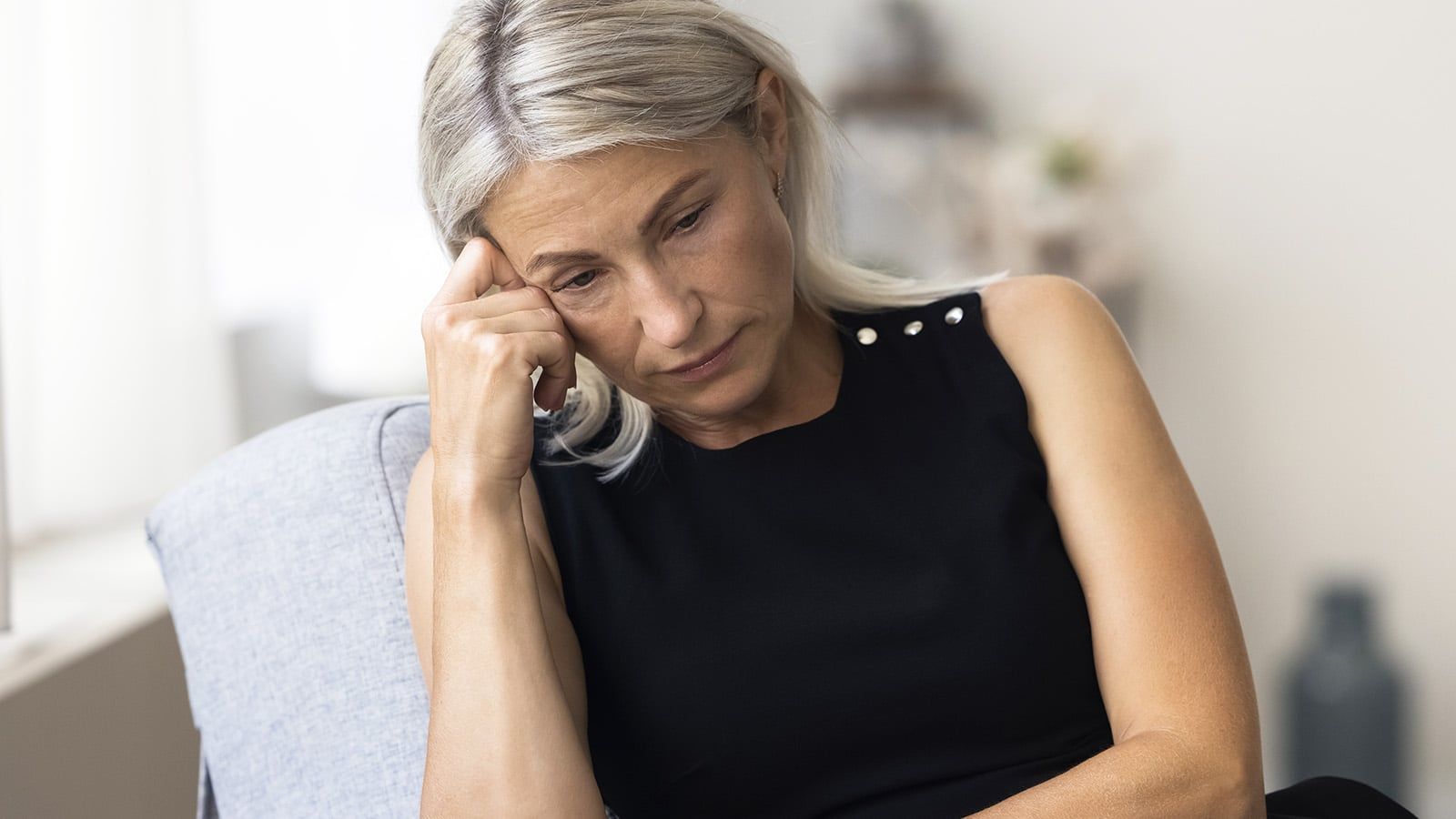 A woman with light hair wearing a black sleeveless top sits on a couch, resting her head on her hand and looking down with a thoughtful or concerned expression.