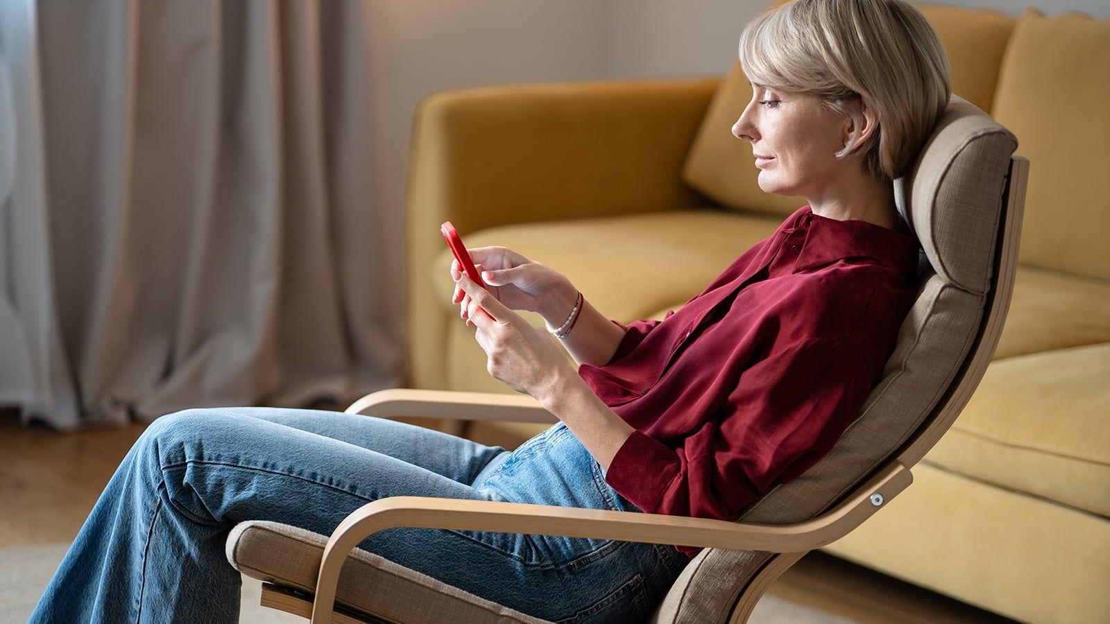 A woman with short blonde hair, wearing a red blouse and blue jeans, sits in a wooden chair and looks at her smartphone—perhaps taking a moment for herself amid the demands of family caregiving. A yellow sofa and curtains are visible in the background.