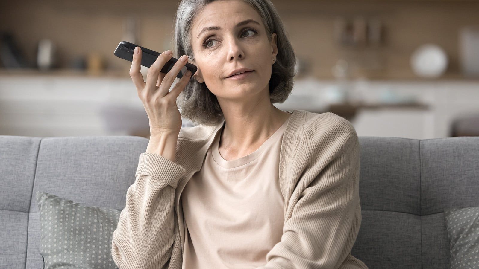 A woman with short gray hair sits on a couch, holding her smartphone to her ear on speaker mode, possibly listening to a psychologist’s voicemail. She wears a beige cardigan and light shirt in a cozy living room setting.