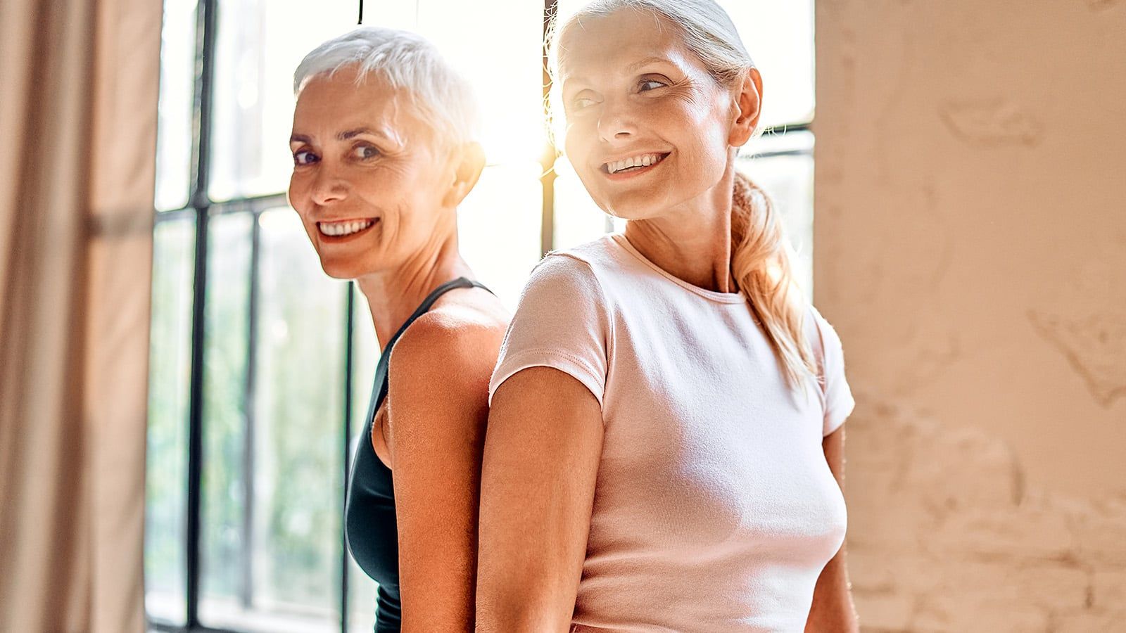 Two smiling older women with light hair stand back to back in front of large windows, wearing casual athletic tops. Sunlight streams in, creating a warm and cheerful atmosphere—perfect for sharing an inspirational quote of the day, just like Jane Fonda would.