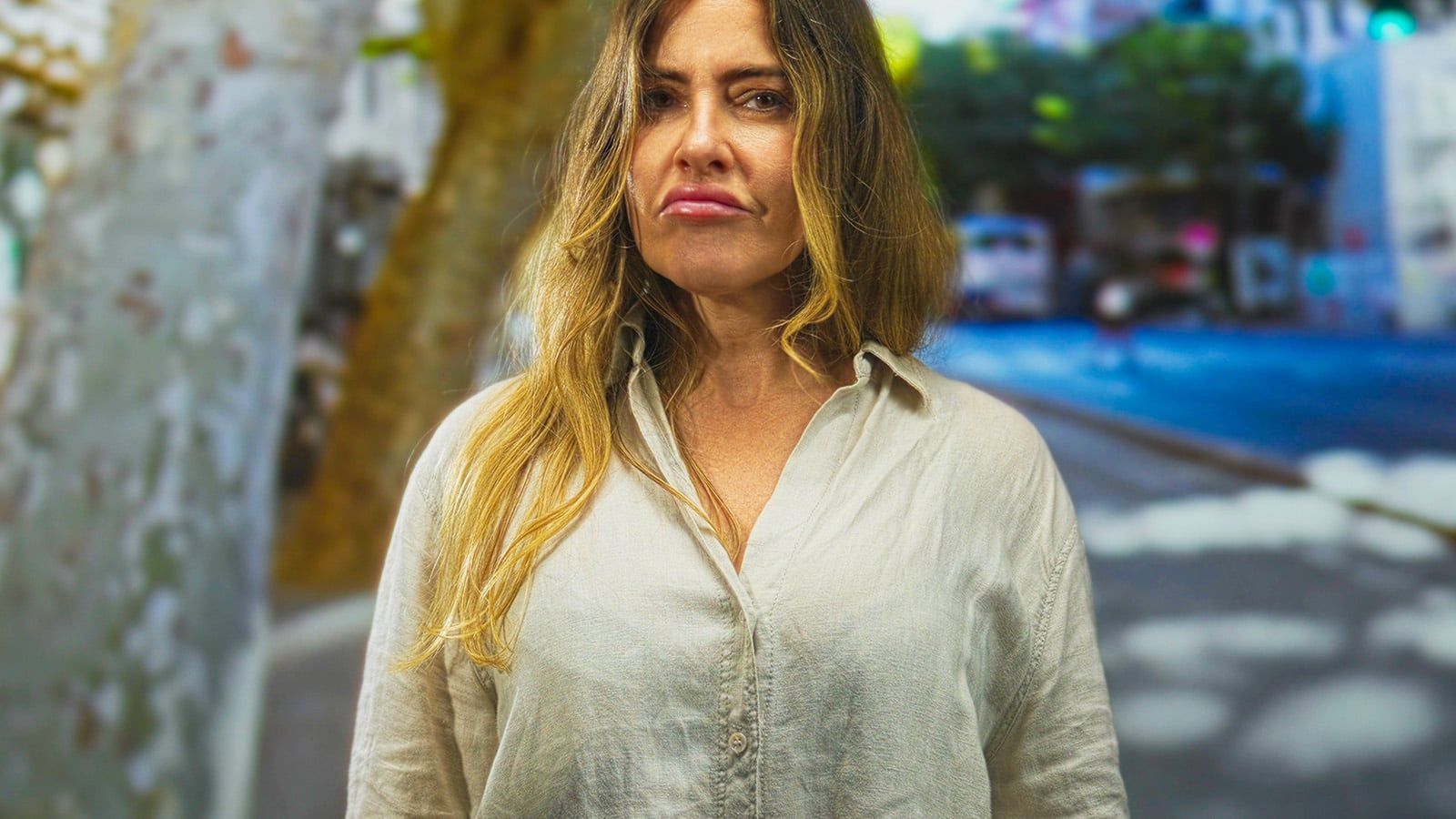 A woman with long, blonde hair wearing a light-colored button-up shirt stands outdoors on a city street, looking directly at the camera with a serious expression. The background is blurred with trees and buildings.