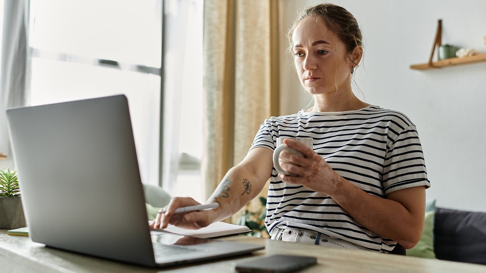 A woman in a striped shirt sits at a table, focused on her laptop with a mug in hand. Surrounded by a notebook, pen, and phone, she reflects the challenges of work-life balance in today’s digital workplace.