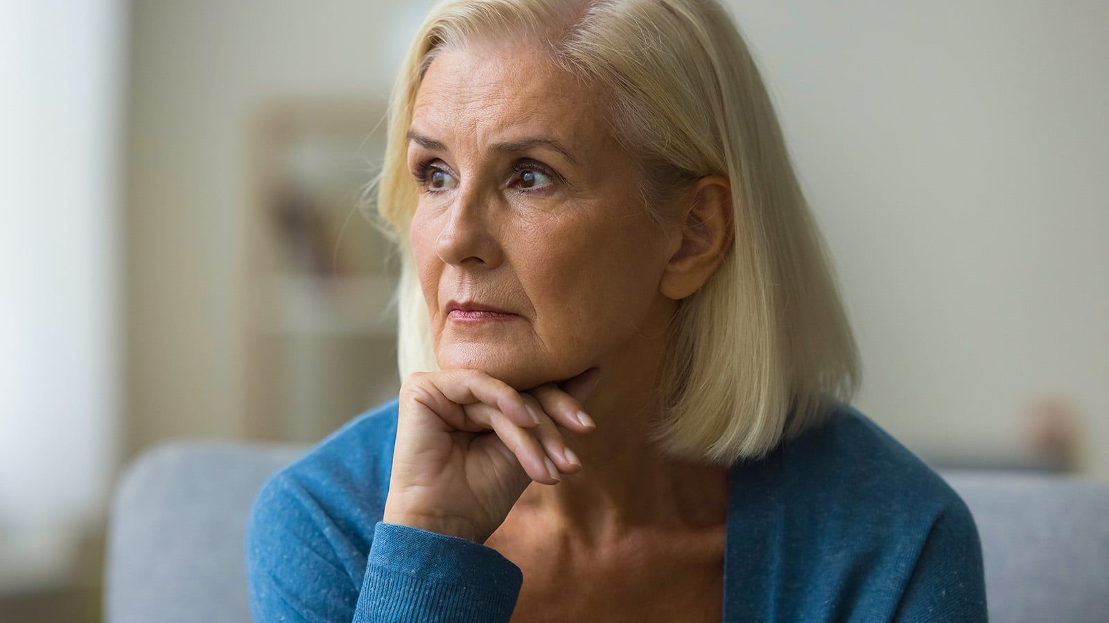 An older woman with short blonde hair sits indoors, resting her chin on her hand and gazing thoughtfully to the side, as if reflecting on regret in old age and worrying about the past, much like findings from a Cornell researcher study.