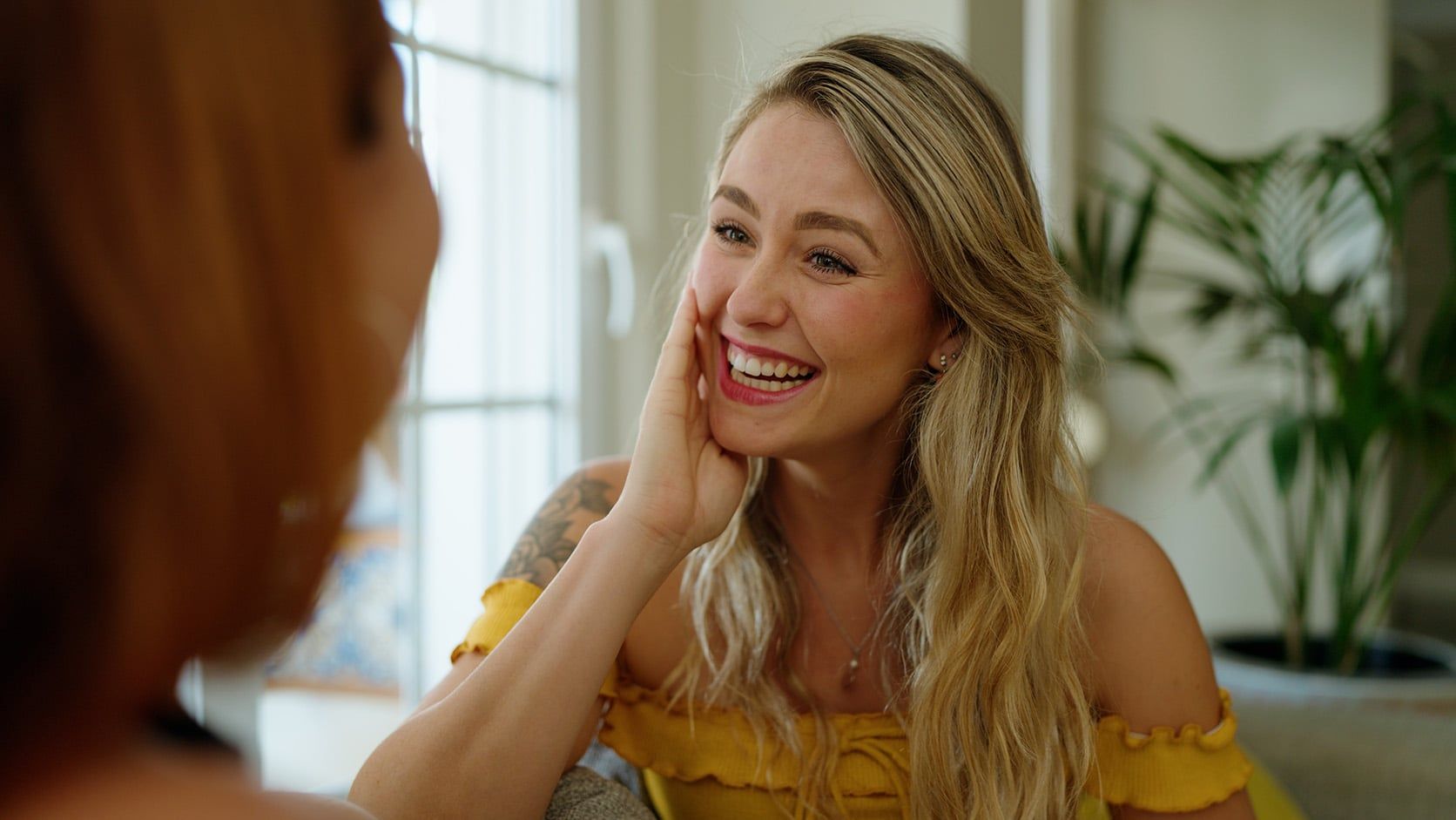 A smiling woman with long blonde hair and a yellow off-shoulder top rests her hand on her face while talking to another person indoors, showing signs of confidence as she appears happy, engaged, and secure in a room filled with sunlight.