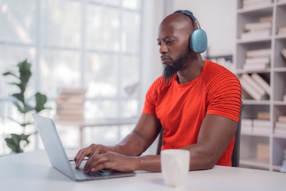 A black man in an orange shirt working on his laptop and listening to music with his headphones