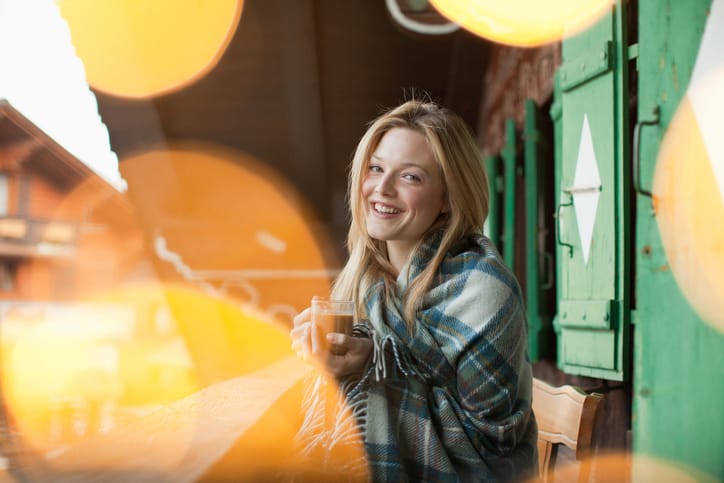 Smiling woman wrapped in a blanket and drinking coffee on cabin porch