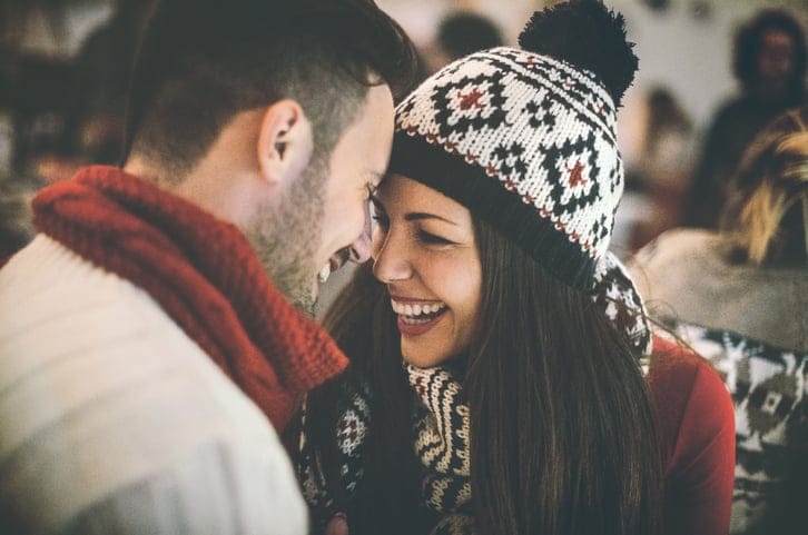 Closeup of happy mid 20's couple having fun at cosy coffee place on winter night. They are sitting face to face by a large window. Both wearing warm casual clothing. Blurry Christmas lights and decoration in background. Tilt shot,toned image.