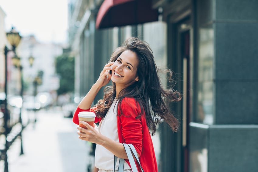 Smiling young woman with phone and coffee cup walking on the street and looking back over her shoulder.