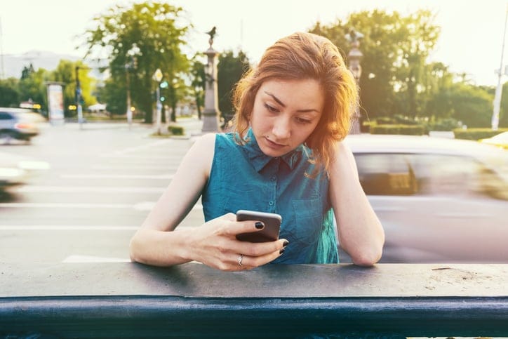 Teenage girl sitting on a sidewalk near the boulevard and looking at her cell phone. Long exposure image with blurred cars and blue dark toning.