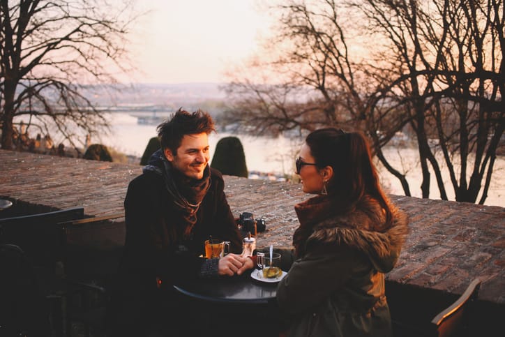 Young couple sitting at the outdoors coffee shop in the park. A couple in love holding hands, drinking tea, and smiling at each other. Young man and young woman facing each other amongst the park trees on a sunny winter afternoon.