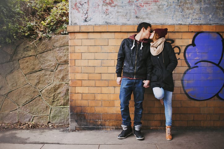 Affectionate teenage couple kissing outdoors against a wall on street. Mixed race couple in love.