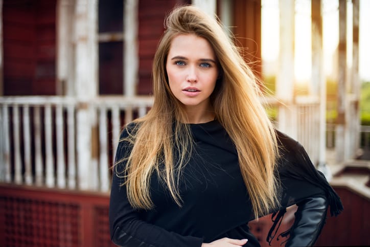 Beautiful blonde girl with long hair posing against old village house at sunset time
