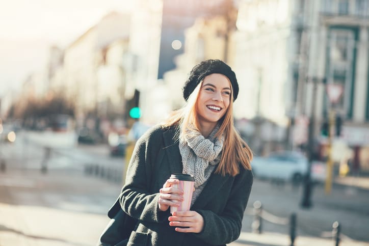Smiling young woman in warm clothing walking under the sunlight at city scene, with copy space