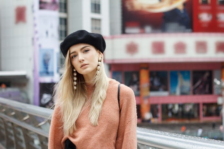 A beautiful young blonde woman dressed in a large warm pink sweater with a black beret and gorgeous earrings. She goes through a big spectacular Chinese city in autumn and enjoying every moment.