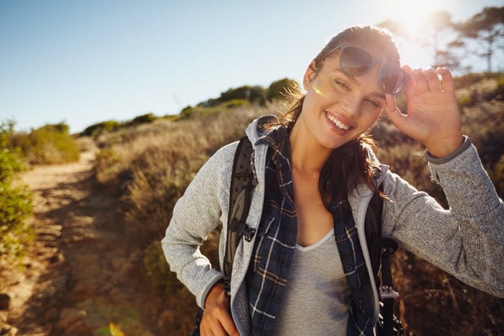 Portrait of a happy young hiker woman in nature. Young caucasian woman wearing sunglasses looking at camera with a backpack on a summer day. Eco tourism and freedom concept