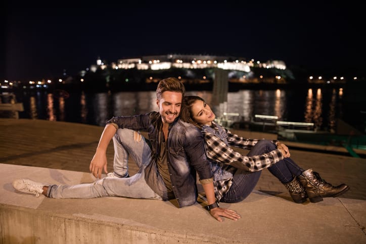 Happy couple sitting back to back on a riverside at night.