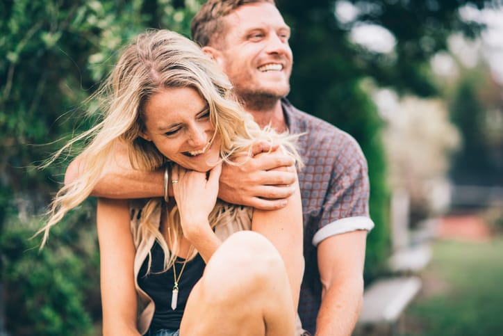 Young couple sitting on a bench in a park on a summer day. Man is hugging his girlfriend. They are smiling, man is looking to the right side of the image, woman is looking down.