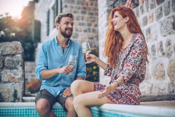 Romantic couple having aperitif near the swimming pool. Sitting at poolside and kissing.