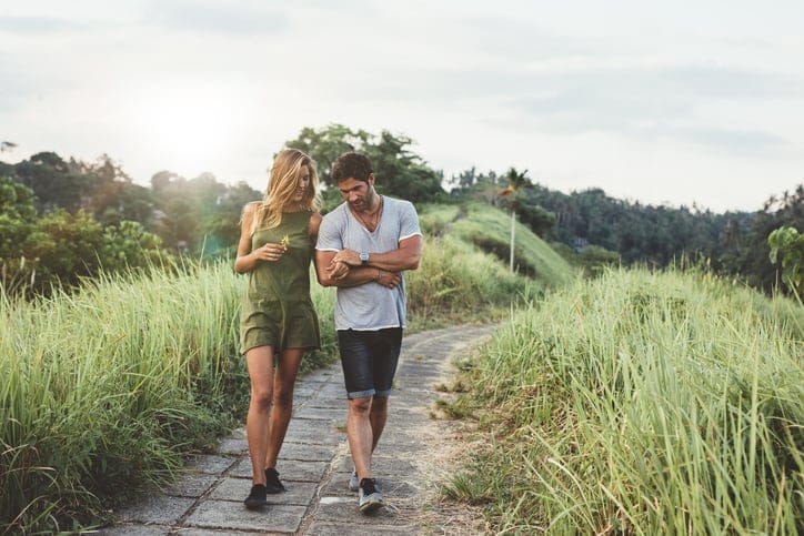 Young couple holding hands and walking through pathway in rural field. Man and woman walking along tall grass field.